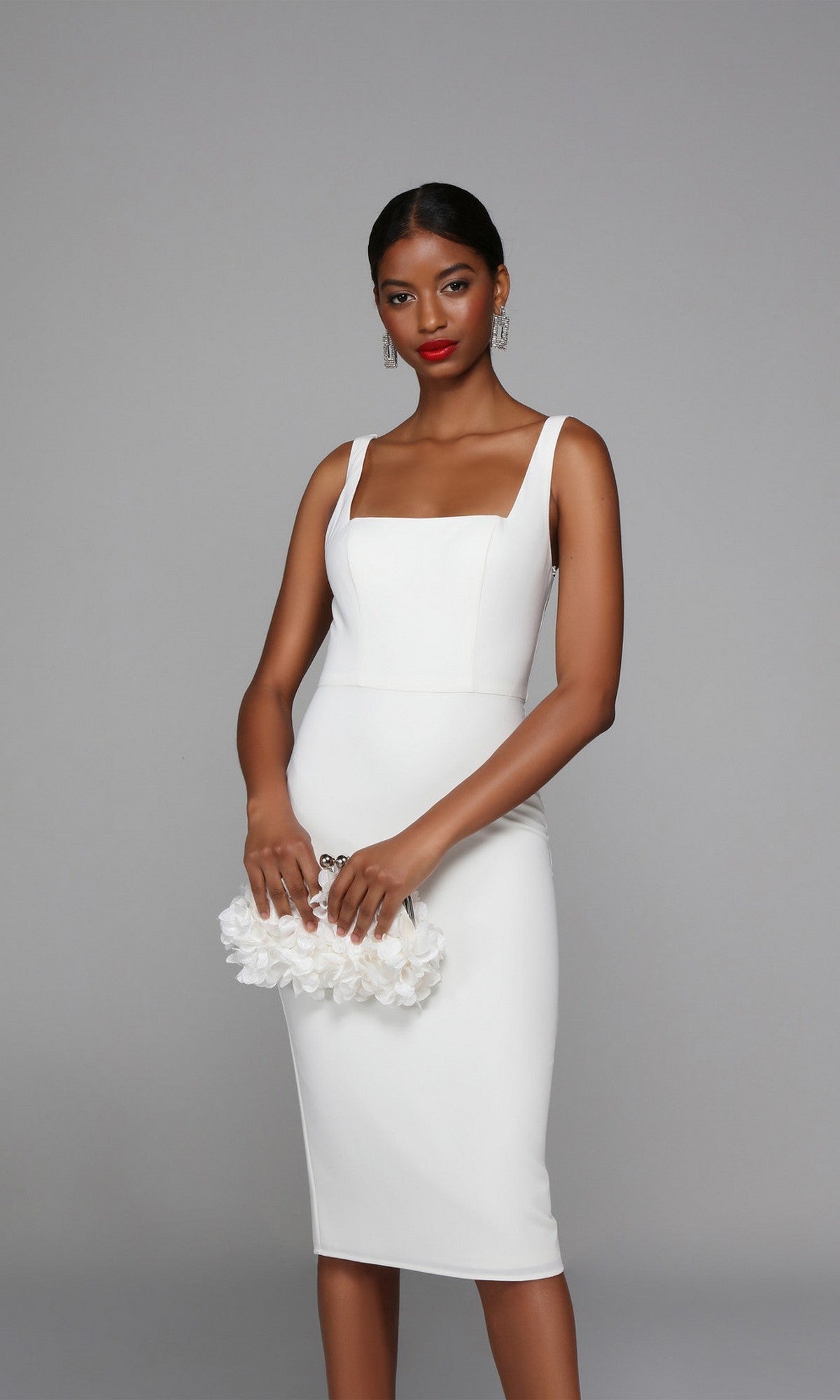 A woman in a fitted white Knee Length Semi-Formal Dress: Alyce 70010 holds a white floral clutch, her hair pulled back and wearing drop earrings, standing against a plain gray background.